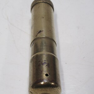 May include: A cylindrical brass object with a ridged top and a darker, metallic section. The body has a worn, aged appearance with a small hole near the base. The object's overall length is approximately 10 cm.