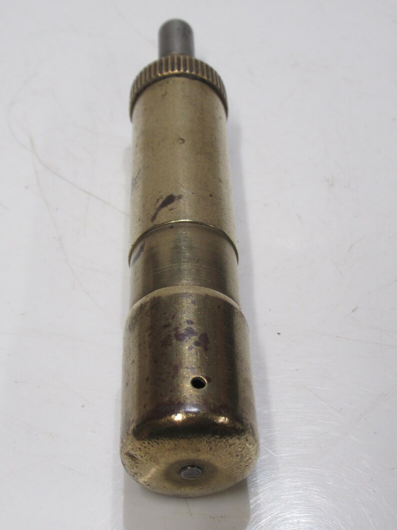 May include: A cylindrical brass object with a ridged top and a darker, metallic section. The body has a worn, aged appearance with a small hole near the base. The object's overall length is approximately 10 cm.