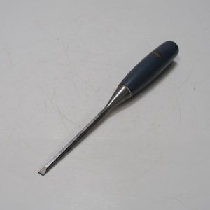 Puede incluir: Un formón para carpintería con un mango azul oscuro y una hoja plateada. El formón está diseñado para cortar y dar forma a la madera con precisión. La herramienta está sobre una superficie blanca.