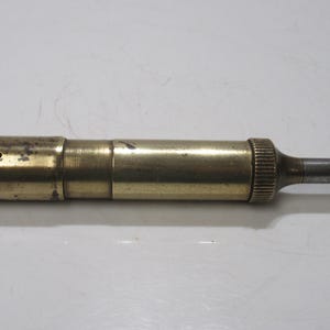 May include: A vintage brass object with a cylindrical body and a small, protruding metal tip. The brass has a tarnished, aged appearance, with a textured, ridged end. The object appears to be a tool or instrument.