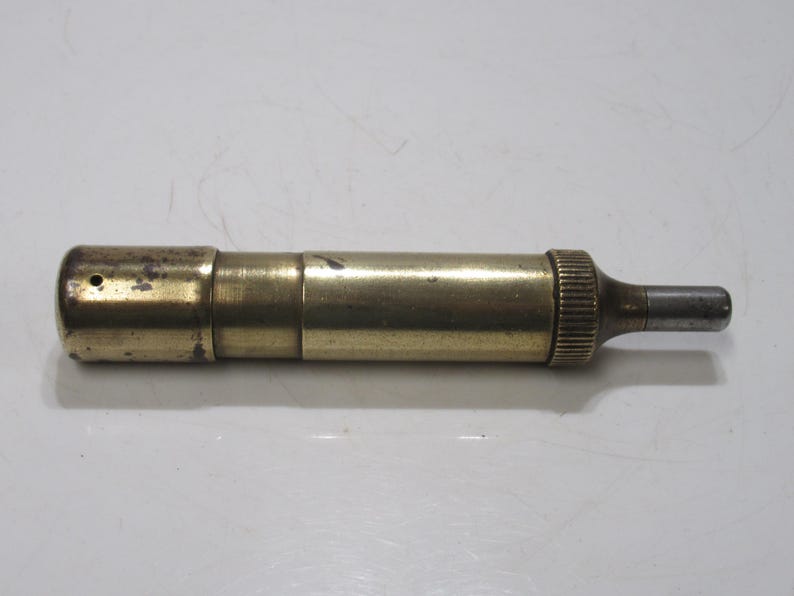 May include: A vintage brass object with a cylindrical body and a small, protruding metal tip. The brass has a tarnished, aged appearance, with a textured, ridged end. The object appears to be a tool or instrument.