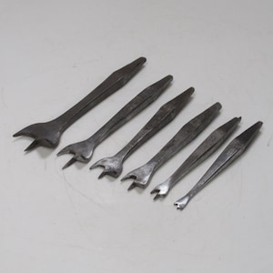 Puede incluir: Conjunto de seis herramientas metálicas antiguas, probablemente para carpintería o trabajos similares. Cada herramienta tiene un mango plano y un extremo bifurcado o curvado. Las herramientas varían en tamaño, con un acabado oscuro y envejecido.