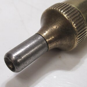May include: Close-up of a vintage brass and steel tool. The tool has a cylindrical body with a ridged top and a tapered, metallic tip. The metal shows signs of age and wear, with a slightly tarnished appearance.