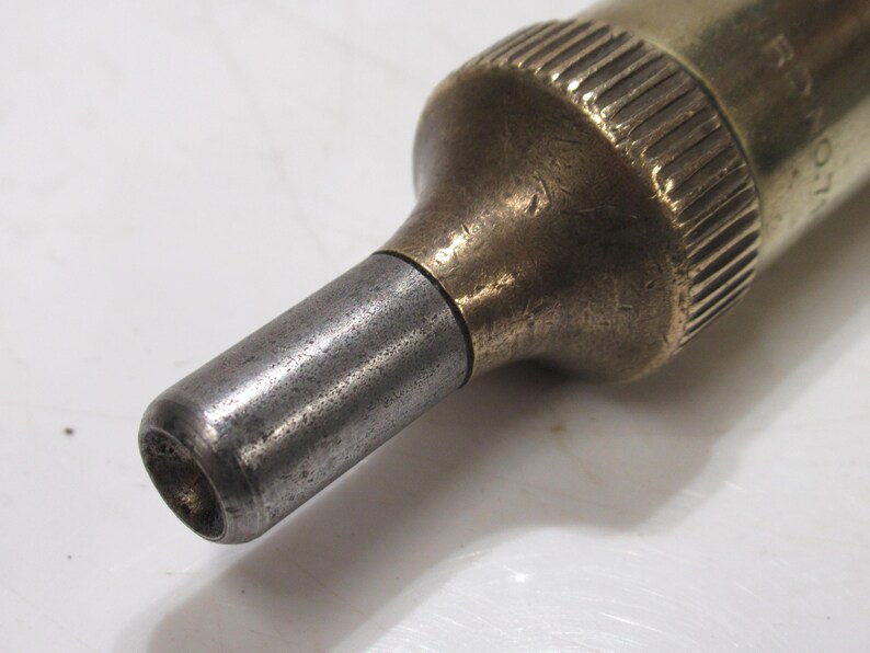 May include: Close-up of a vintage brass and steel tool. The tool has a cylindrical body with a ridged top and a tapered, metallic tip. The metal shows signs of age and wear, with a slightly tarnished appearance.