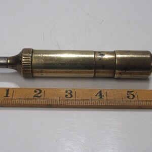 May include: A vintage brass tool with a cylindrical body and a small, metallic tip. The tool is approximately 13 cm long and lies next to a wooden ruler. The tool has a ribbed section near the tip.
