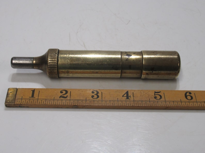May include: A vintage brass tool with a cylindrical body and a small, metallic tip. The tool is approximately 13 cm long and lies next to a wooden ruler. The tool has a ribbed section near the tip.
