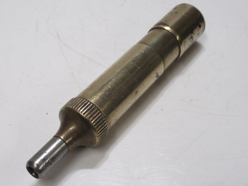 May include: A vintage brass and steel tool with a cylindrical body. The tool has a ribbed grip section and a tapered nozzle. The metal has a worn, aged appearance.
