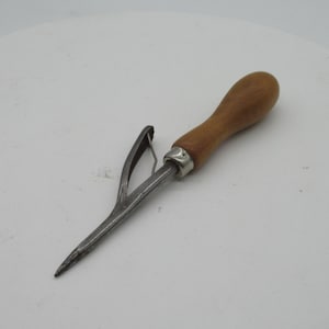 May include: A vintage metal tool with a wooden handle. The tool has a pointed end and a curved, pronged section. The handle is a light brown color, and the metal appears to be silver. The tool is likely used for a specific craft or trade.