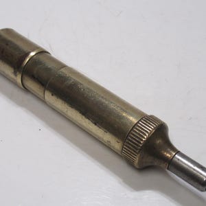 May include: A vintage brass tool with a cylindrical body and a tapered end. The tool has a series of rings and ridges for grip and a silver-coloured tip. The brass has a slightly aged patina.