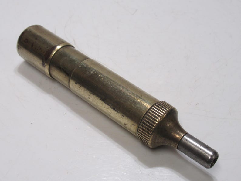 May include: A vintage brass tool with a cylindrical body and a tapered end. The tool has a series of rings and ridges for grip and a silver-coloured tip. The brass has a slightly aged patina.