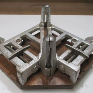 May include: A silver-colored corner clamp tool with adjustable screws, mounted on a dark brown wooden base. The clamp is designed to hold pieces at a 90-degree angle, ideal for woodworking projects.