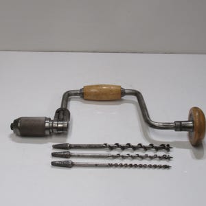 Puede incluir: Un taladro manual antiguo con mango de madera y tres brocas. El taladro tiene un marco de metal con un mango de madera y un pomo de madera. Las brocas son de metal y tienen un diseño en espiral.