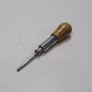 May include: A vintage screwdriver with a wooden handle and a metal shaft. The handle is a light brown color, and the metal shaft is silver. The screwdriver is designed for slotted screws and is a hand tool.