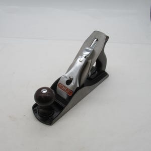 Peut inclure: Un rabot à main Stanley, avec une base noire, une lame argentée et un bouton en bois brun foncé. Le mot "STANLEY" est visible. Cet outil de menuiserie est conçu pour lisser et façonner les surfaces en bois.