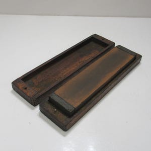 May include: A wooden sharpening stone with a dark brown, rectangular stone set in a wooden base. The lid is open, revealing the stone and its wooden case. The wood has a rich, aged appearance.