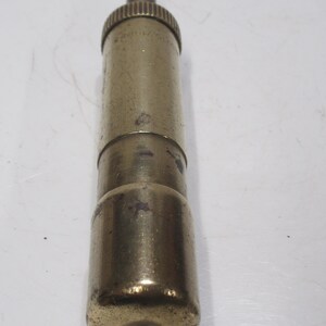 May include: A vintage brass-coloured cylindrical object with a textured top and a rounded base. The object has a small, dark-coloured rod extending from the top. The surface shows signs of wear, suggesting age and use. The item appears to be a tool or a component of a larger device.