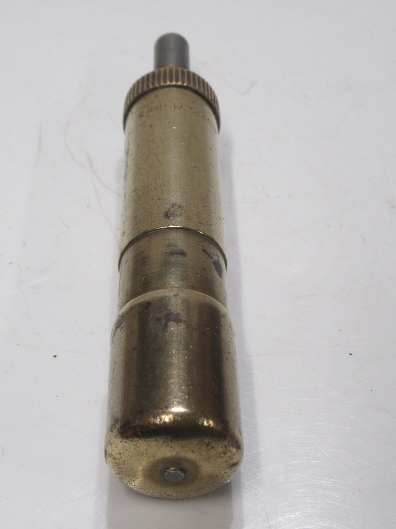 May include: A vintage brass-coloured cylindrical object with a textured top and a rounded base. The object has a small, dark-coloured rod extending from the top. The surface shows signs of wear, suggesting age and use. The item appears to be a tool or a component of a larger device.