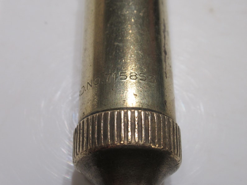 May include: Close-up of a vintage brass object with the text "D.NO. 715853" engraved on its surface. The object has a ridged base and a slightly tarnished appearance, suggesting age and use.