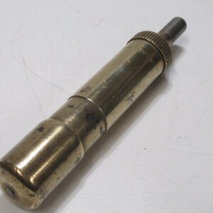 May include: A vintage brass object with a cylindrical body and a textured, ridged top. The object has a tarnished appearance, suggesting age. The design includes a smaller, darker metal piece extending from the top. The item is likely a tool or a decorative piece.