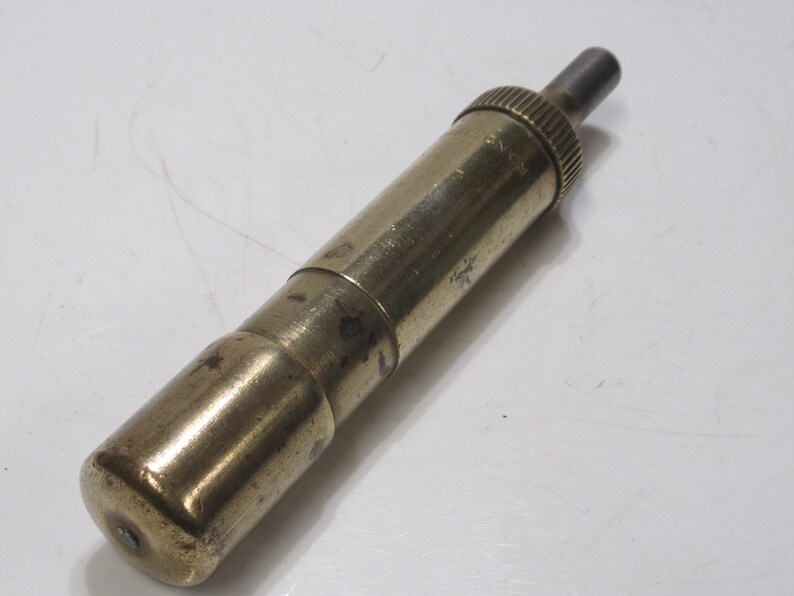 May include: A vintage brass object with a cylindrical body and a textured, ridged top. The object has a tarnished appearance, suggesting age. The design includes a smaller, darker metal piece extending from the top. The item is likely a tool or a decorative piece.