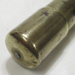 May include: Close-up of a brass object with a rounded end. The brass has a worn, aged appearance with some darker spots. The object appears to be cylindrical, possibly a part of a larger mechanism or tool.