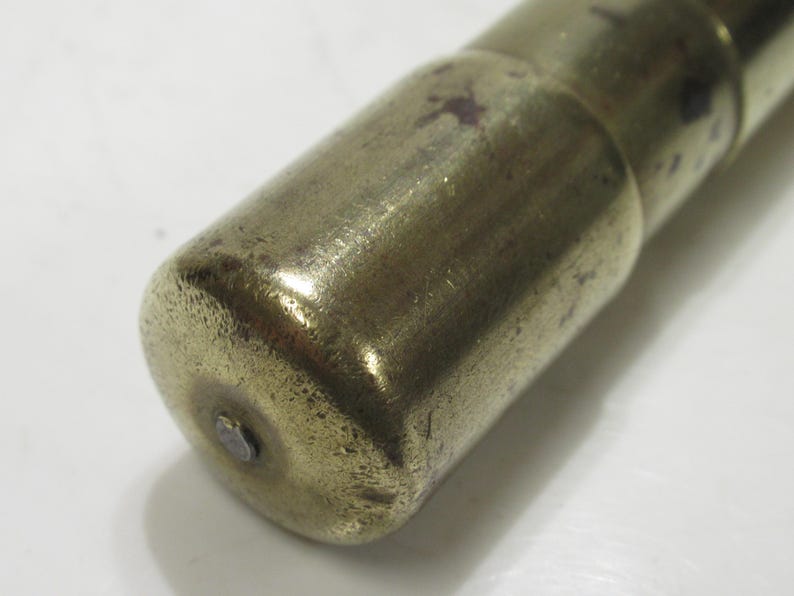 May include: Close-up of a brass object with a rounded end. The brass has a worn, aged appearance with some darker spots. The object appears to be cylindrical, possibly a part of a larger mechanism or tool.