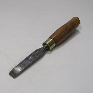 May include: A woodworking chisel with a wooden handle, brass ferrule, and a steel blade. The handle is a light brown color, and the blade is a dark gray. The chisel is designed for shaping and cutting wood.