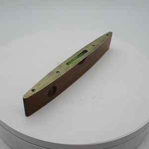 Puede incluir: Un nivel de madera vintage con detalles de latón. El nivel es rectangular, con latón en la parte superior e inferior. Cuenta con un vial transparente que contiene una burbuja de líquido verde para medir el nivel. Probablemente utilizado en la construcción o carpintería.