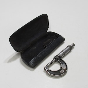 May include: A vintage micrometer and its black leather case are shown on a white surface. The micrometer is made of metal and has a C-shaped frame. The case is open, revealing a dark interior.