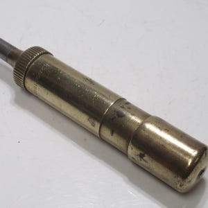 May include: A vintage brass tool with a cylindrical body and a knurled grip. The tool has a dark metal tip and a rounded end. The brass has a tarnished appearance, suggesting age and use. The tool is likely used for a specific mechanical purpose.