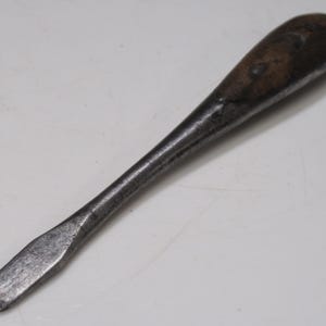 May include: A vintage flathead screwdriver with a dark wooden handle and a metal shaft. The tool is designed for turning slotted screws and shows signs of wear, indicating its age and use. The screwdriver is a hand tool.