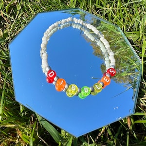 Puede incluir: Un brazalete de cuentas blancas con la palabra "DIBBLES" escrita en cuentas de colores. Las cuentas son rojas, naranjas, amarillas, verdes y azules.