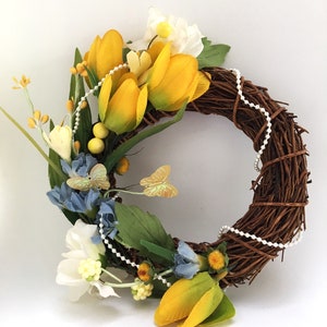 Könnte beinhalten: Ein brauner Zweigkranz, der mit gelben und blauen Blumen, weißen Blumen und Perlen verziert ist.
