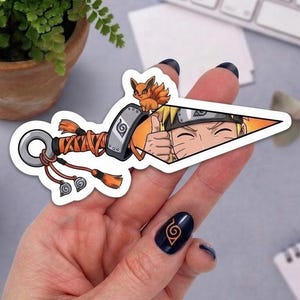 May include: A die-cut vinyl sticker shaped like a kunai knife featuring a cartoon character with blonde hair and a small orange fox. The handle is wrapped in orange fabric with tassels, and the blade displays a character giving a thumbs up.