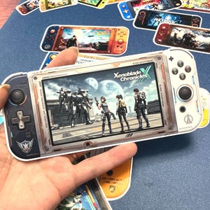 May include: A white and gray handheld gaming console with a screen displaying a video game scene. The console has buttons and a directional pad. Several game console stickers are scattered around on a blue surface.