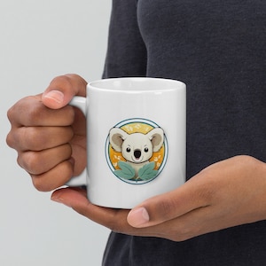 May include: White ceramic mug with a cartoon koala in a circle surrounded by green leaves and a yellow background. The koala has a light brown nose and black eyes.