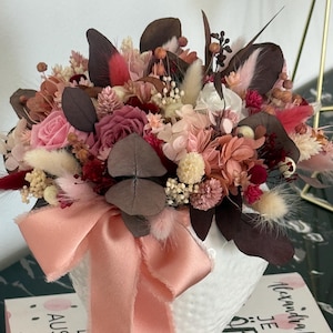 Op de afbeelding: Een bloemstuk in een witte vaas met een perzikkleurig lint. Het boeket bevat rozen, droogbloemen en donkerbruine bladeren. Het arrangement is een mix van roze, perzik en bruine tinten, wat een zachte en elegante uitstraling creëert.