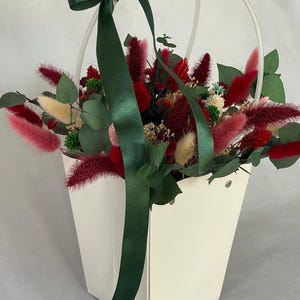 Könnte beinhalten: Eine weiße Papiertüte mit einem grünen Band und einem Strauß getrockneter Blumen. Die Blumen sind rot, rosa und weiß. Die Tasche hat einen Griff.