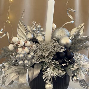 Könnte beinhalten: Eine schwarze Keramikvase mit einem silbernen und weißen Weihnachts-Mittelstück. Das Mittelstück enthält eine weiße Kerze, silberne Kugeln, weiße Beeren und silbernes und schwarzes Laub.