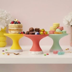 May include: Three colorful cake stands in yellow, coral, and mint green, each with a unique design. The yellow stand holds a small cake topped with berries, the coral stand has chocolates and fruit, and the mint stand displays macarons.