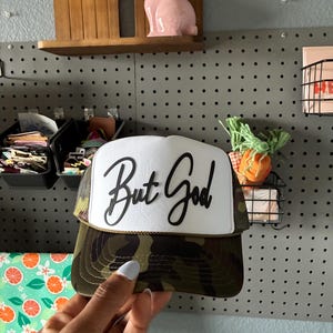 May include: A camouflage trucker hat with a white front panel featuring the script "But God" in black. The hat has a mesh back and a curved brim. The hat is being held up in front of a pegboard.