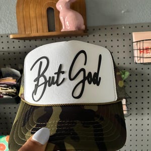May include: A camouflage trucker hat with a white front panel featuring the phrase "But God" in black script. The hat has a curved brim and a mesh back. A gold-colored rope trim accents the brim.