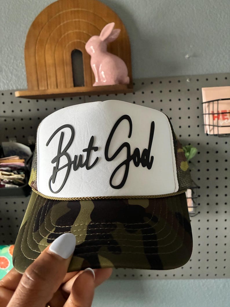 May include: A camouflage trucker hat with a white front panel featuring the phrase "But God" in black script. The hat has a curved brim and a mesh back. A gold-colored rope trim accents the brim.