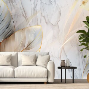 May include: A white couch with two pillows sits in front of a wall with a large abstract print of gold and white leaves on a marble background. A small potted plant sits on a black side table.