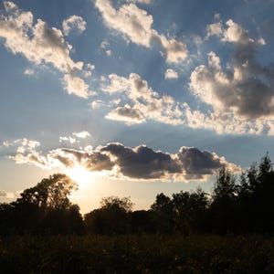 Könnte beinhalten: Ein Sonnenuntergangshimmel mit einer hellen Sonne, die teilweise von dunklen Wolken verdeckt wird und Lichtstrahlen über eine blaue Fläche mit verstreuten weißen Wolken wirft, über einer Silhouette aus dunkelgrünen Bäumen und einem Feld.