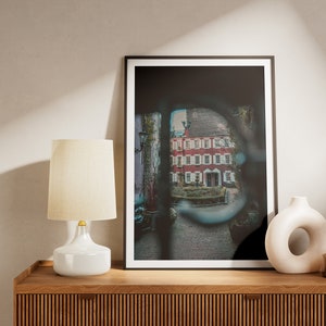 Peut inclure: Une impression encadrée d'un bâtiment en briques rouges avec des garnitures blanches, vue à travers une ouverture arquée. Une lampe blanche avec un abat-jour beige et un vase décoratif sont sur un meuble en bois. Une petite bouteille en verre marron avec des fleurs séchées est également présente.