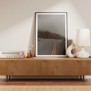 May include: A wooden media console with a slatted front, featuring a vintage stereo receiver, a framed photograph of a winding road through a foggy landscape, a white ceramic lamp with a white shade, and a white ceramic vase with a circular cutout.