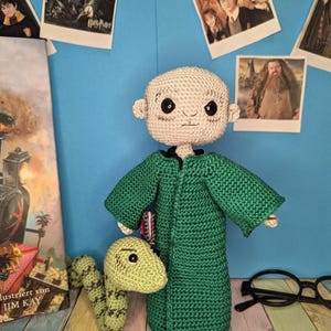 May include: A handmade crochet doll featuring a bald, pale-skinned figure with large black eyes, wearing a long-sleeved green robe. Beside the doll is a small, light green crocheted snake with dark green markings and black eyes.