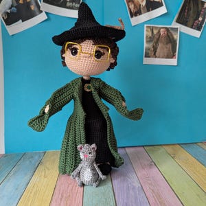 Op de afbeelding: Handgemaakte gehaakte pop van een personage met een zwarte hoed, bril en groene jas, met een kleine grijze gehaakte kat. De pop staat voor een blauwe achtergrond met foto's van filmkarakters.