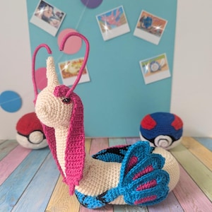 Può includere: Un Milotic all'uncinetto fatto a mano, un personaggio dei Pokémon, in crema, rosa e blu. Il peluche ha un corpo lungo e serpentino con una coda a ventaglio e due lunghe antenne curve. Sono visibili anche due peluche Poké Ball.